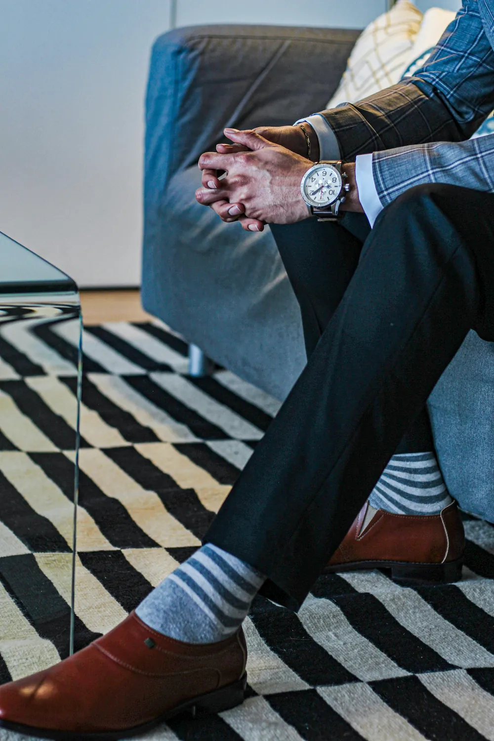 A seated man in dress pants and brown leather shoes wears gray striped socks, with his hands clasped near his knee.