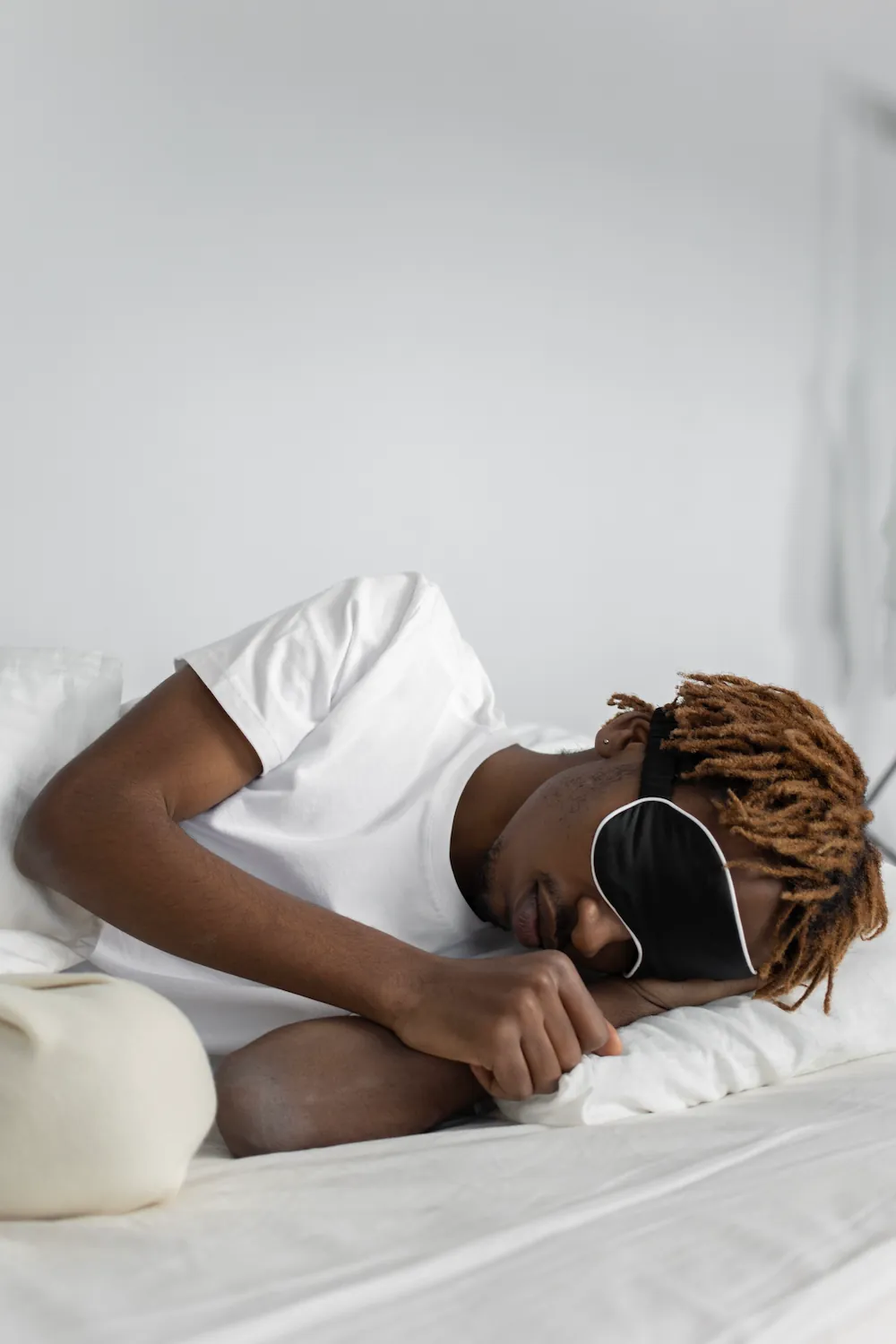 A man sleeps on his side in a white bed wearing a black contoured sleep mask with white trim.