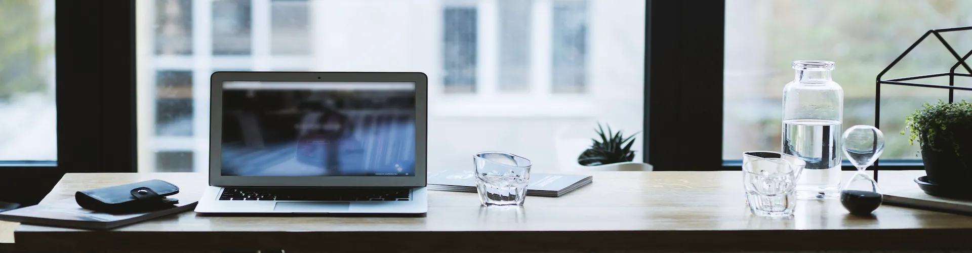 A laptop sits open on a wood desk near tall windows, alongside a wallet, glasses of water, and a small hourglass.