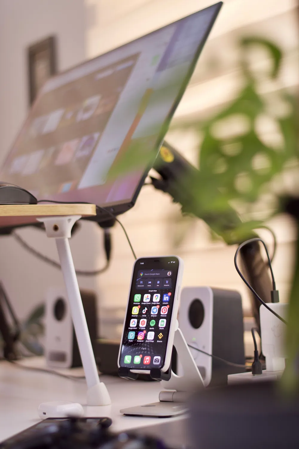A laptop tilts upward on a raised stand beside a smartphone propped in its own stand, with small speakers and a potted plant on the desk.