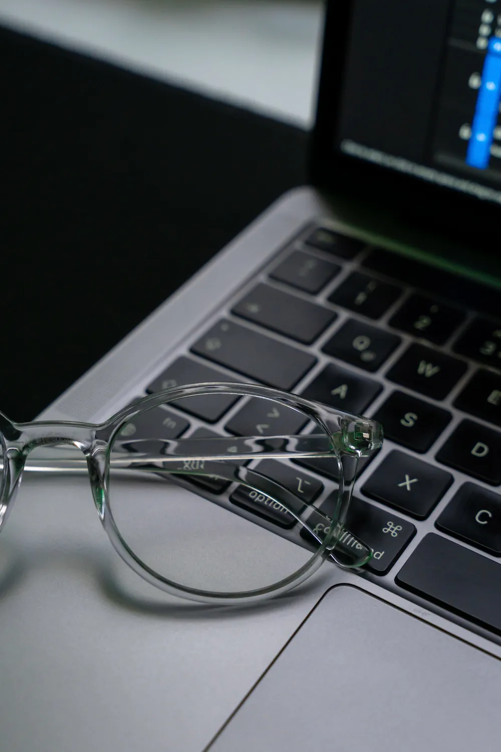 A pair of clear-frame glasses rests on a laptop keyboard with a colorful bar graph glowing on the screen behind them.