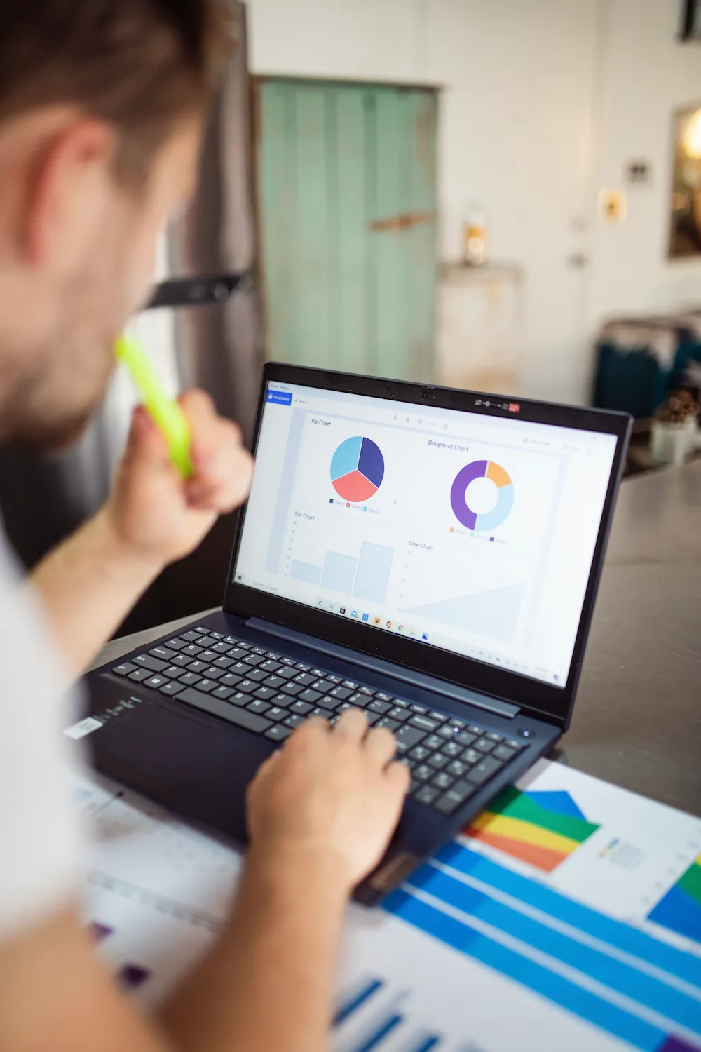 A man reviews a laptop screen showing pie, doughnut, and bar charts while holding a highlighter, with printed color charts spread on the desk.
