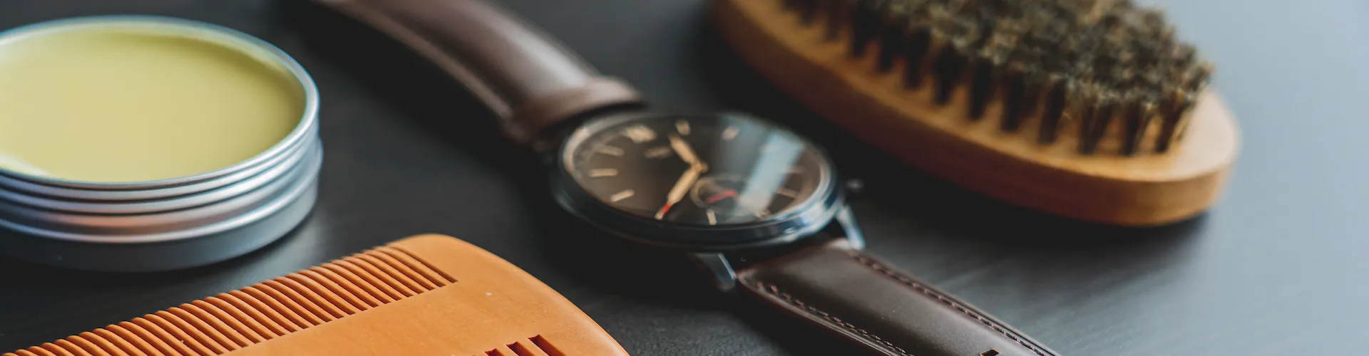 A grooming flat lay on a dark surface with a tin of balm, a wooden comb, a wristwatch, and a boar bristle brush.