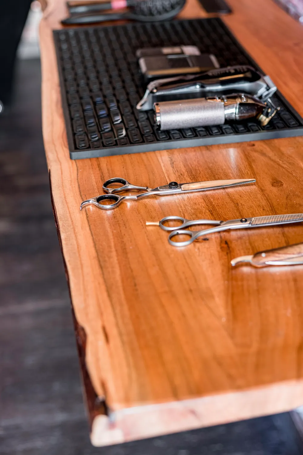 A pair of grooming scissors and thinning shears rest on a wooden counter with barber tools blurred in the background.