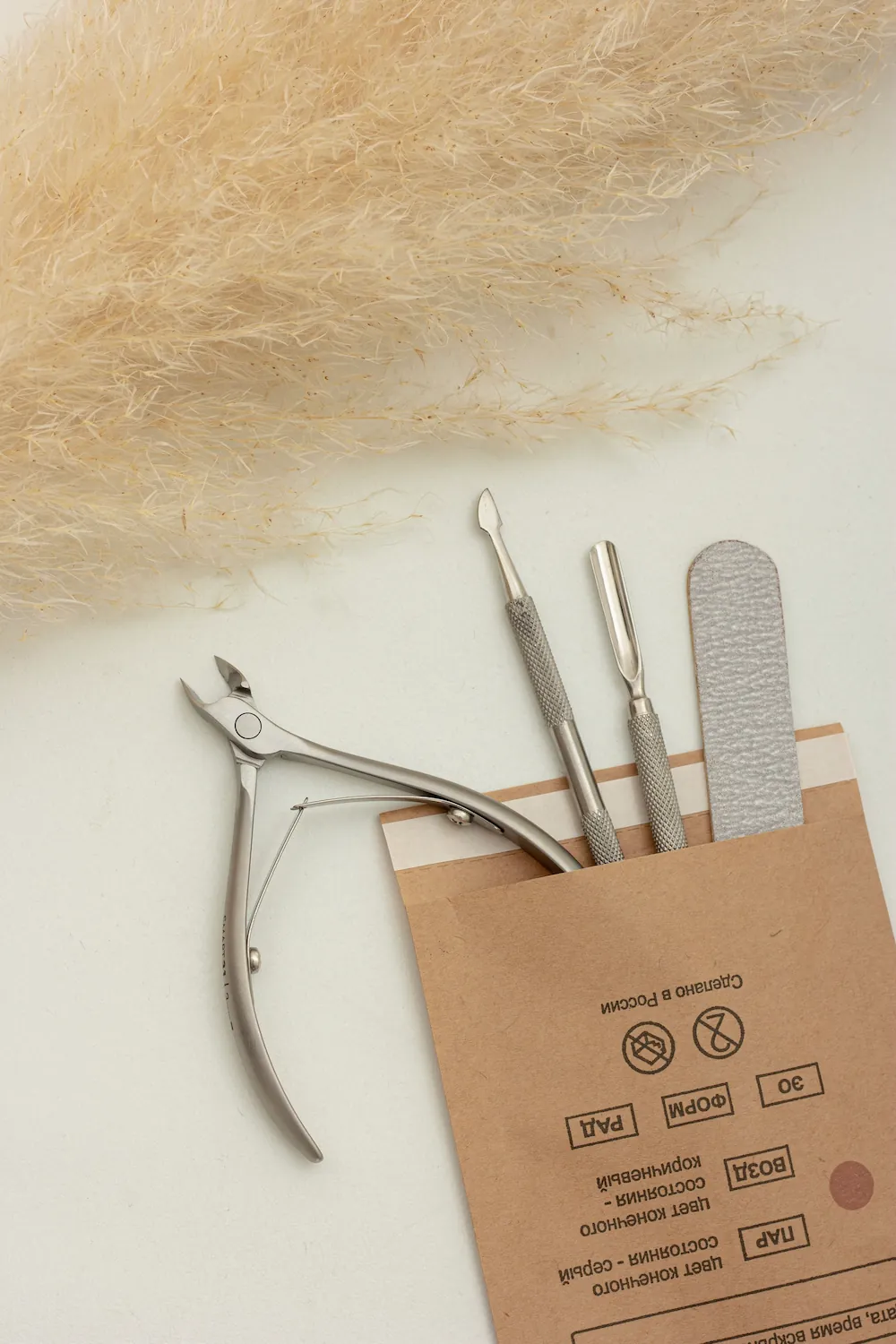 A precision nail clipper, a set of cuticle tools, and a nail file rest beside kraft paper packaging, with dried pampas grass in the background.