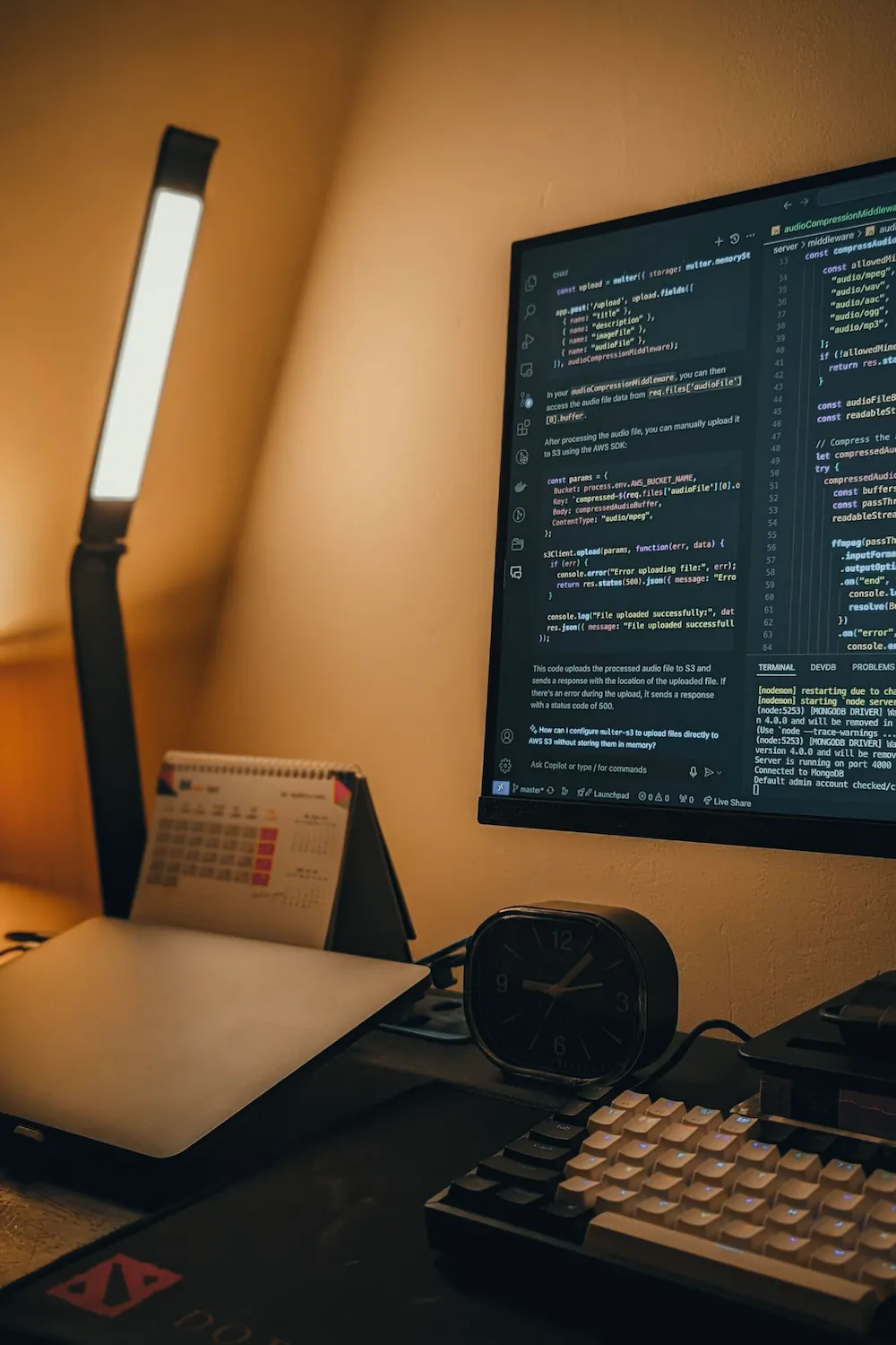 An illuminated desk lamp glows warmly beside a monitor, a small calendar, a clock, and a keyboard in a dim room.