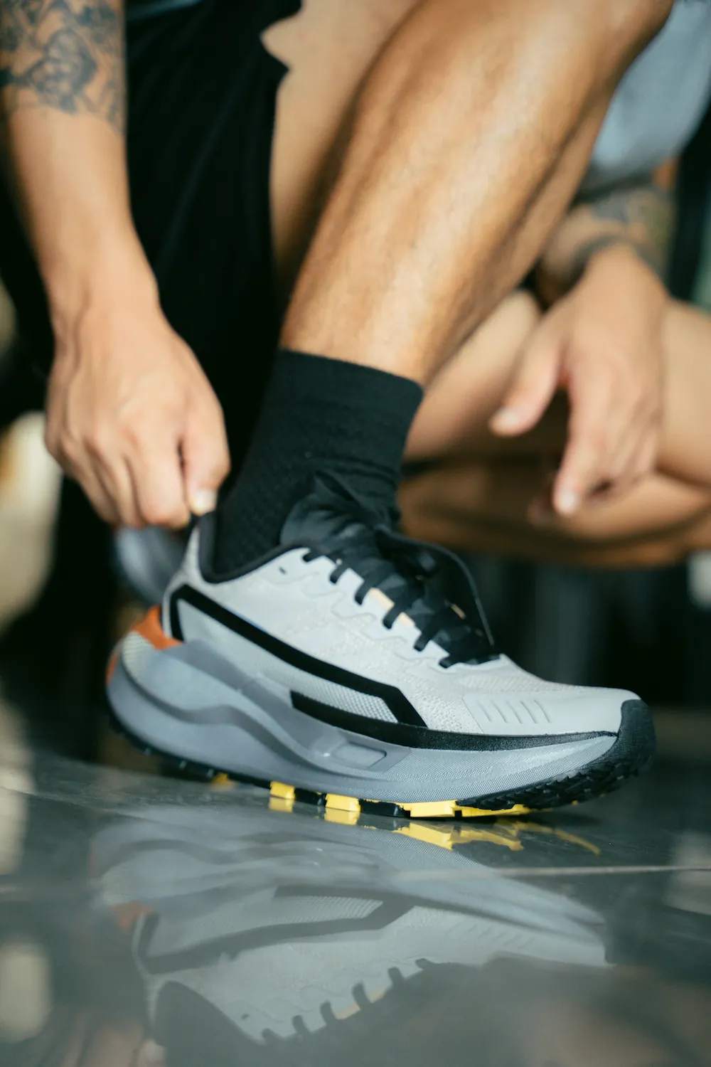 A man ties the laces of a gray and black running shoe while wearing black socks, with tattoos visible on his forearm and calf.