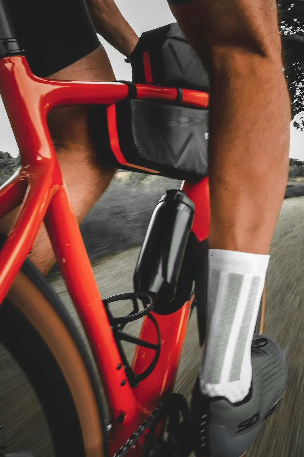 A close-up of a cyclist riding a red road bike with a black frame bag and saddle bag attached, wearing white socks and cycling shoes.