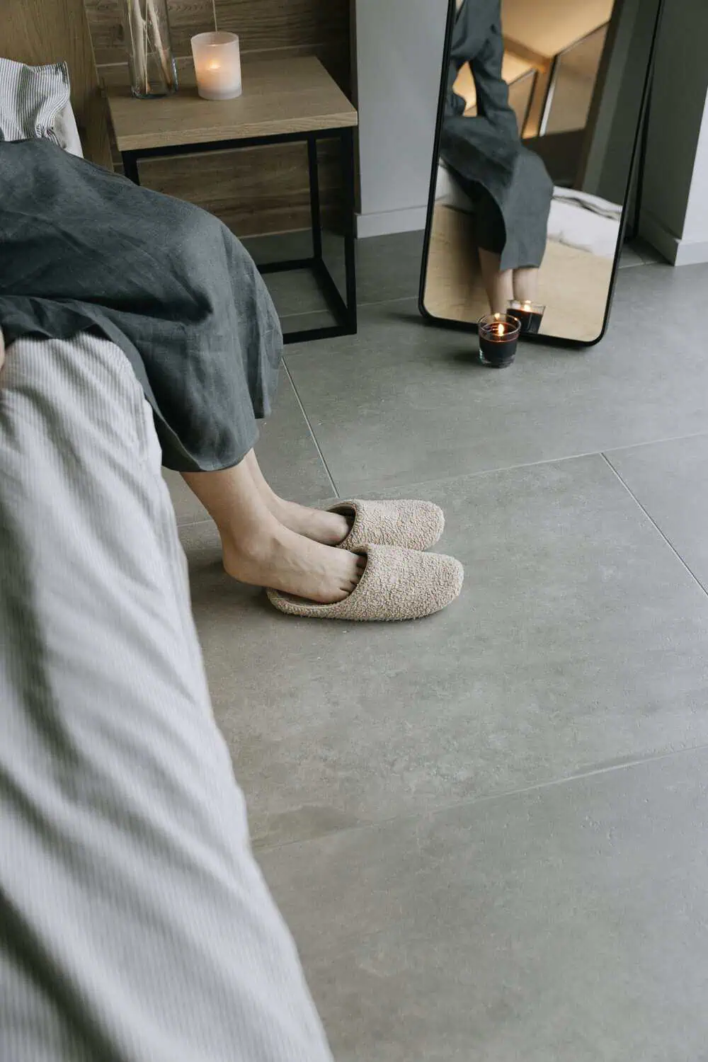 A pair of beige teddy-fabric slipper mules sit on a concrete floor beside a bed, with a lit candle and a full-length mirror reflecting the room in the background.