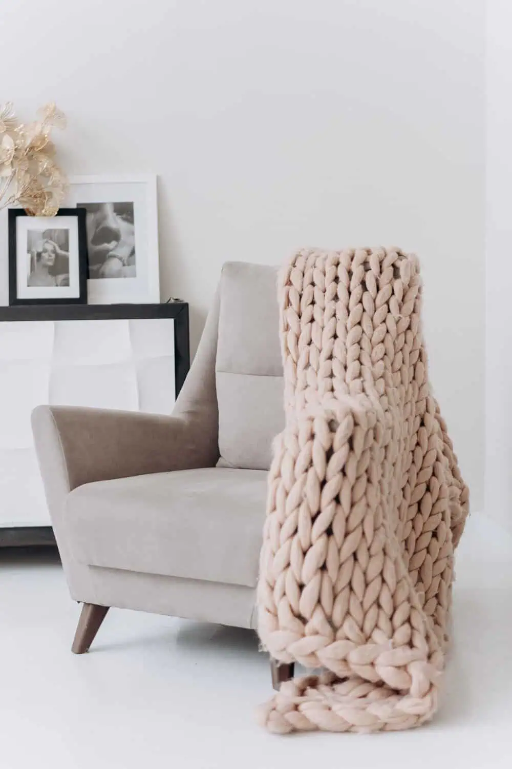 A thick, chunky-knit blush pink throw blanket drapes over the arm of a gray velvet chair in a minimalist living room.