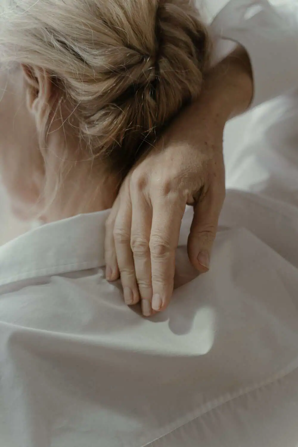 A close-up of a woman's hand resting on the back of her neck and shoulder while wearing a loose white linen top, with her hair pulled into a low bun.
