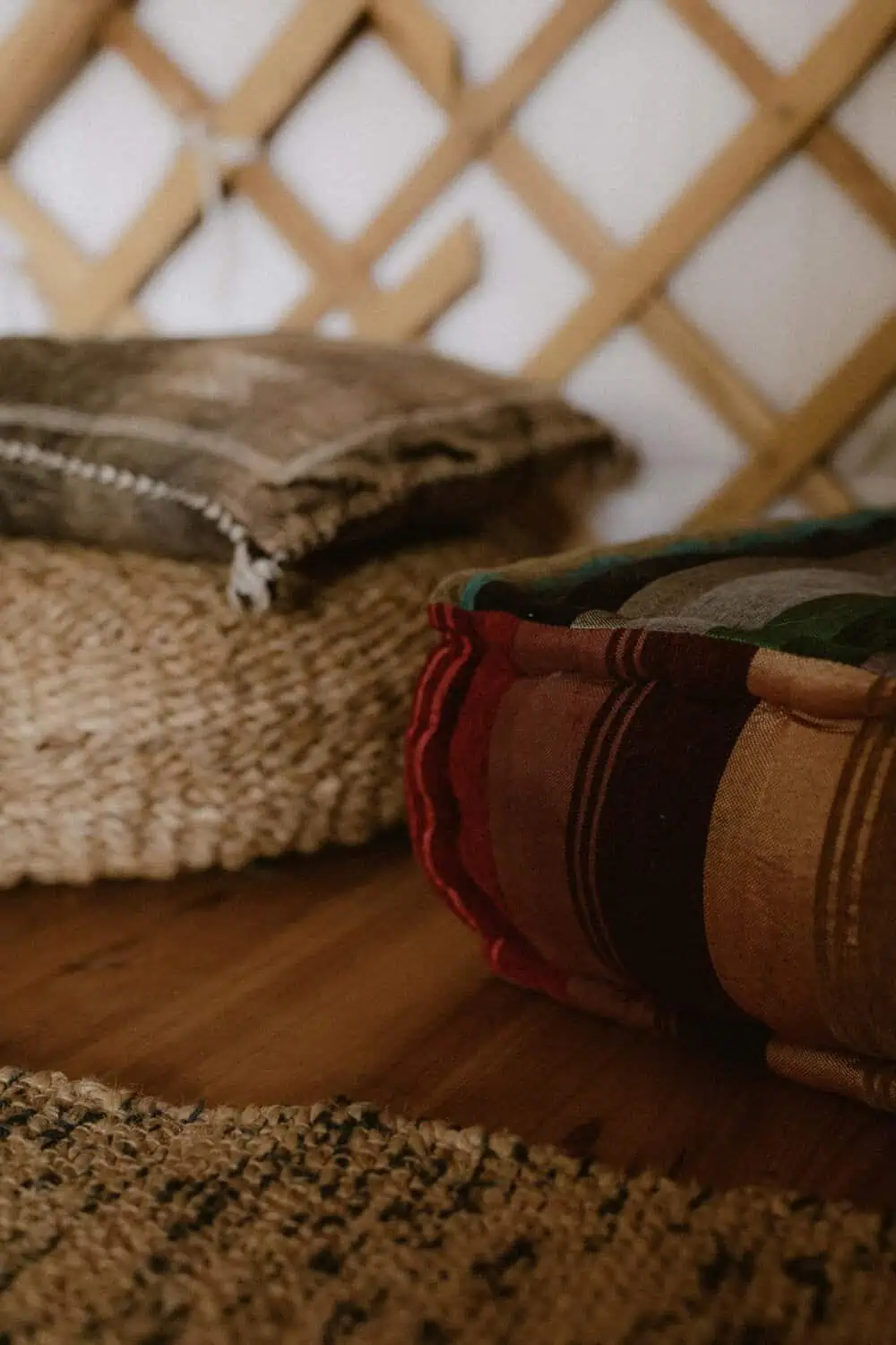 Two round floor cushions, one in a muted plaid fabric and one on a woven jute base, sit on a wood floor next to a textured area rug.
