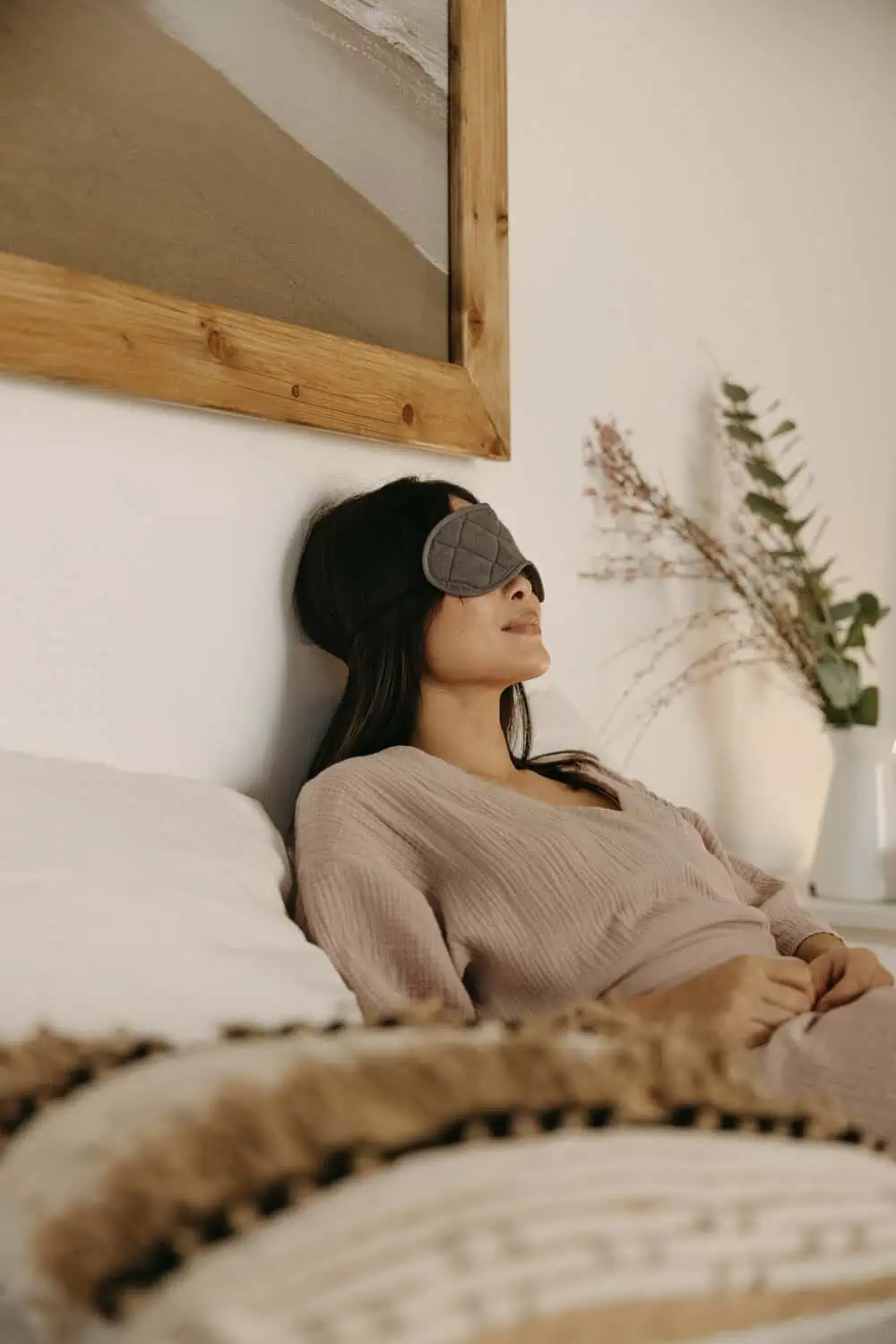 A woman reclines against a headboard with a gray quilted eye mask covering her eyes, wearing a soft knit sweater with a vase of dried branches beside the bed.