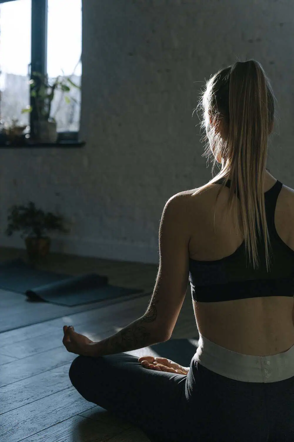 A woman sits cross-legged on the floor of a dim, softly lit room, meditating with her back to the camera and a small potted plant on the windowsill nearby.