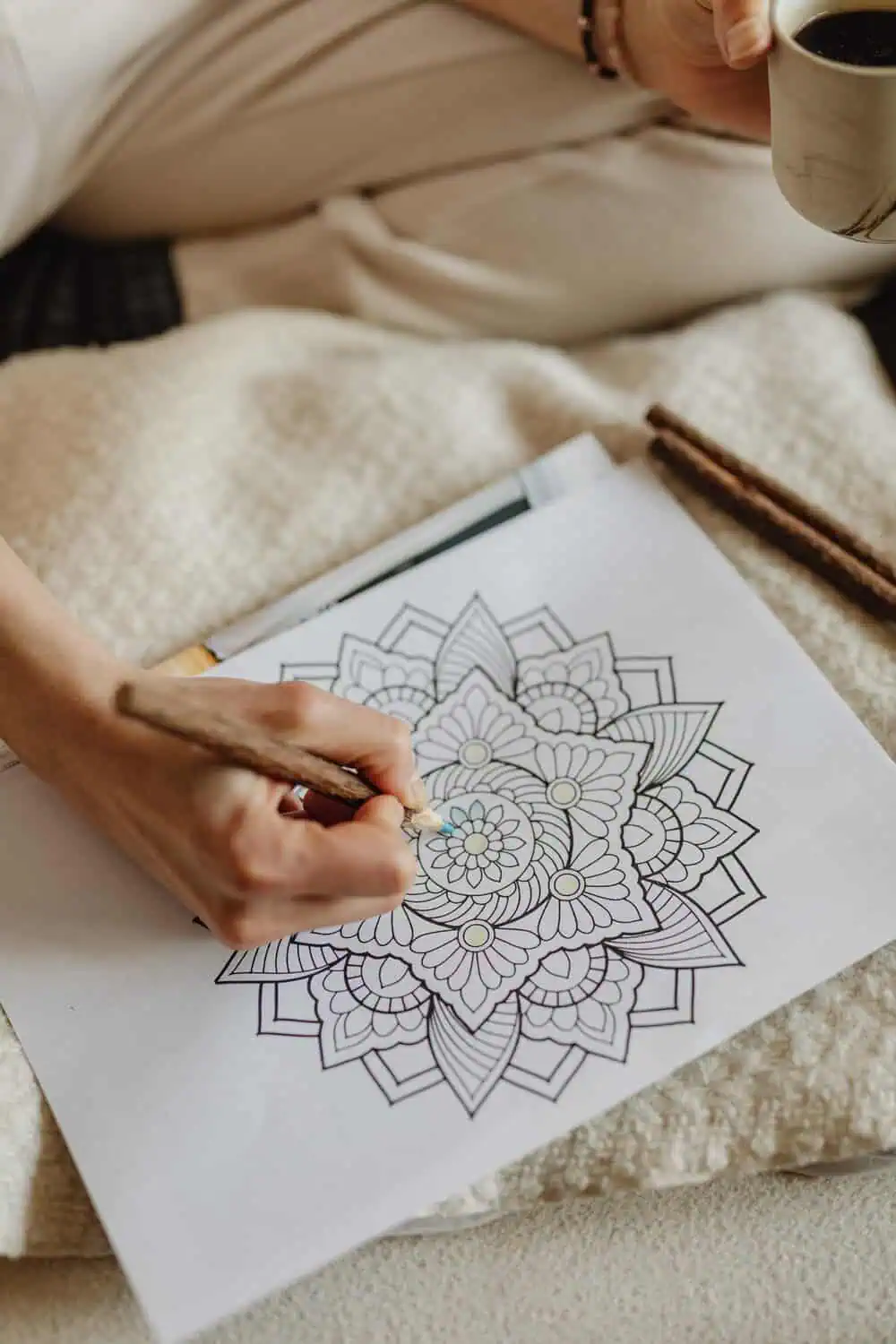 A hand holds a colored pencil while filling in a floral mandala coloring page, with a cup of coffee held in the other hand and colored pencils resting nearby on a cozy blanket.