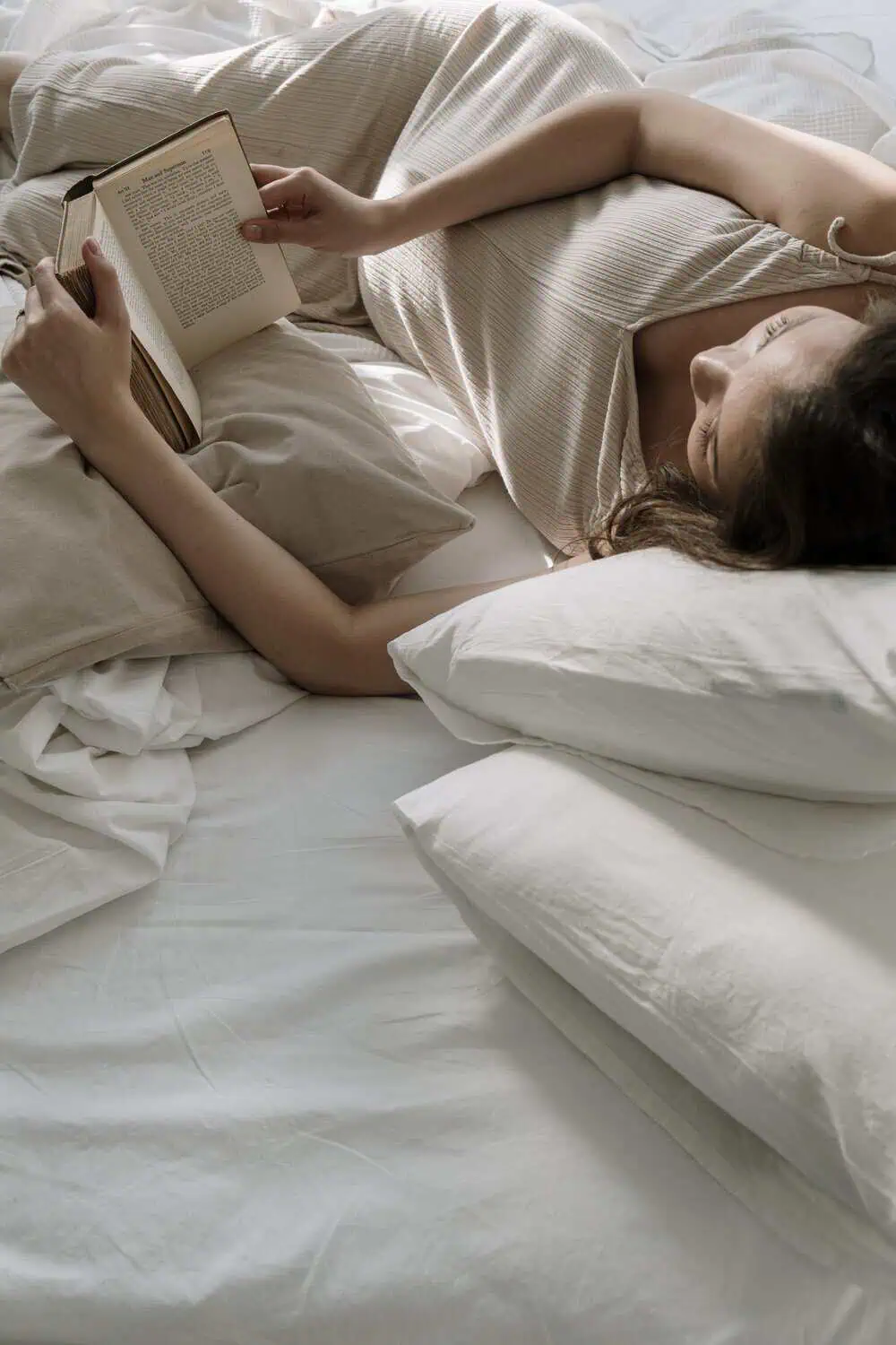 A woman lies in bed reading a paperback book, propped up on several pillows with soft natural light coming through the room.