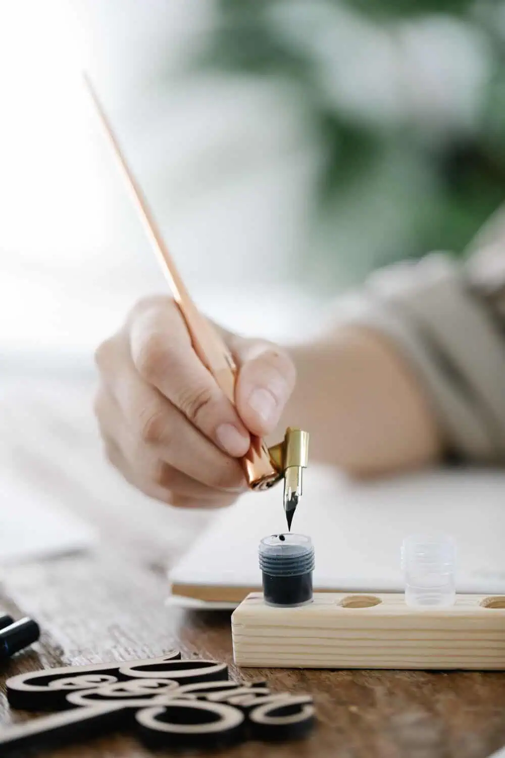 A hand dips a rose gold calligraphy pen into a small jar of black ink at a wooden workstation, with cut wooden script lettering pieces resting nearby.