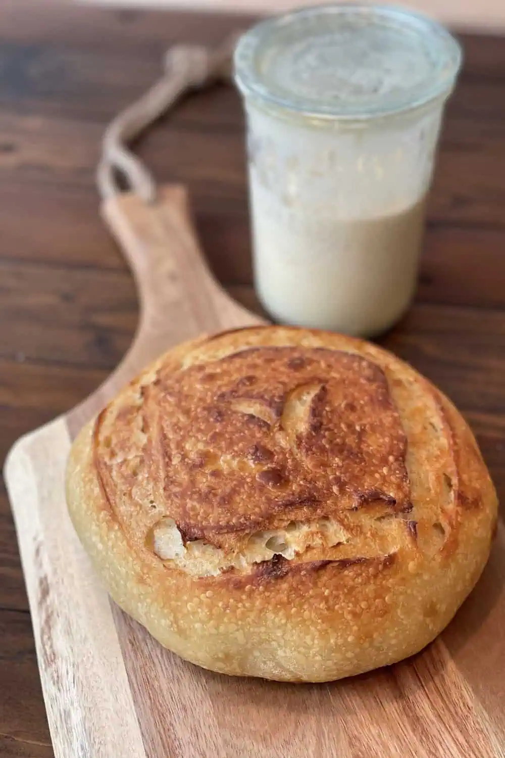 A round, golden-crusted sourdough loaf sits on a wooden board in front of a glass jar of active sourdough starter.
