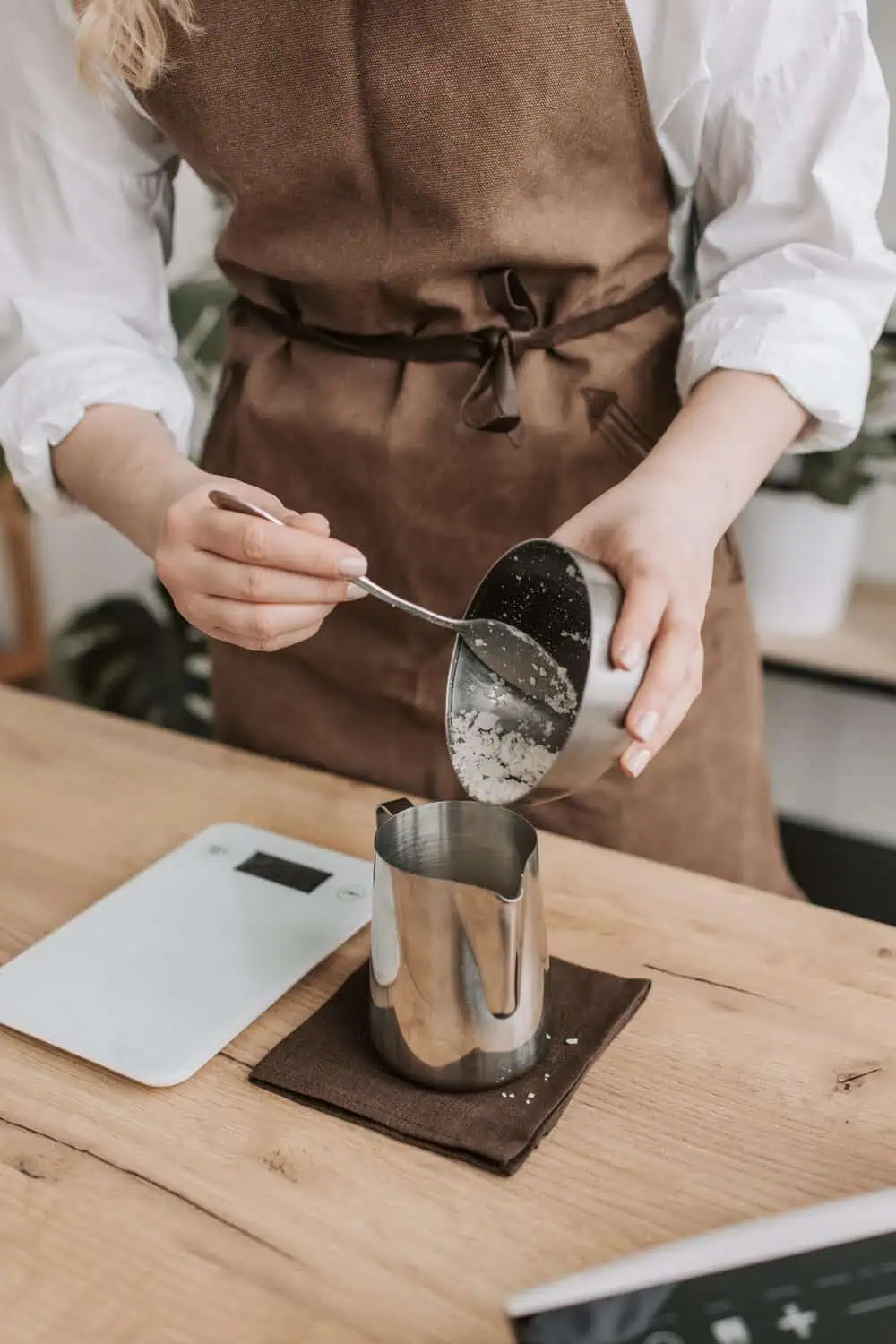 A person in an apron spoons flour from a metal cup into a steel pitcher resting on a slim white digital kitchen scale on a wood table.