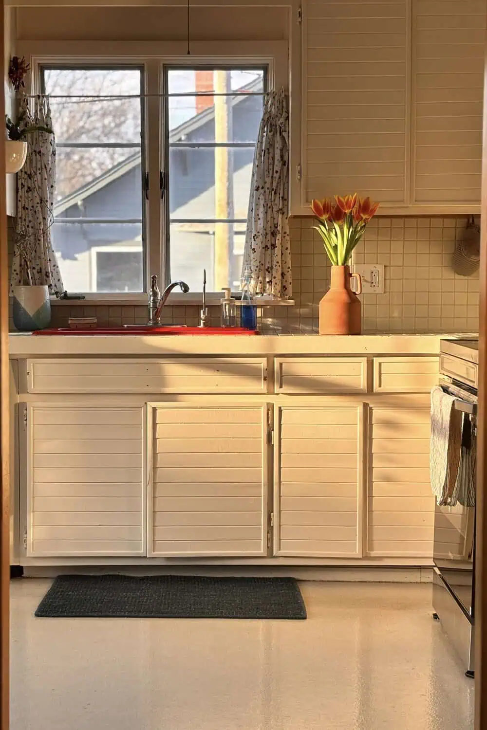 A dark cushioned floor mat sits in front of a vintage-style kitchen sink with a red basin insert, striped curtains, and a vase of orange tulips on the counter.