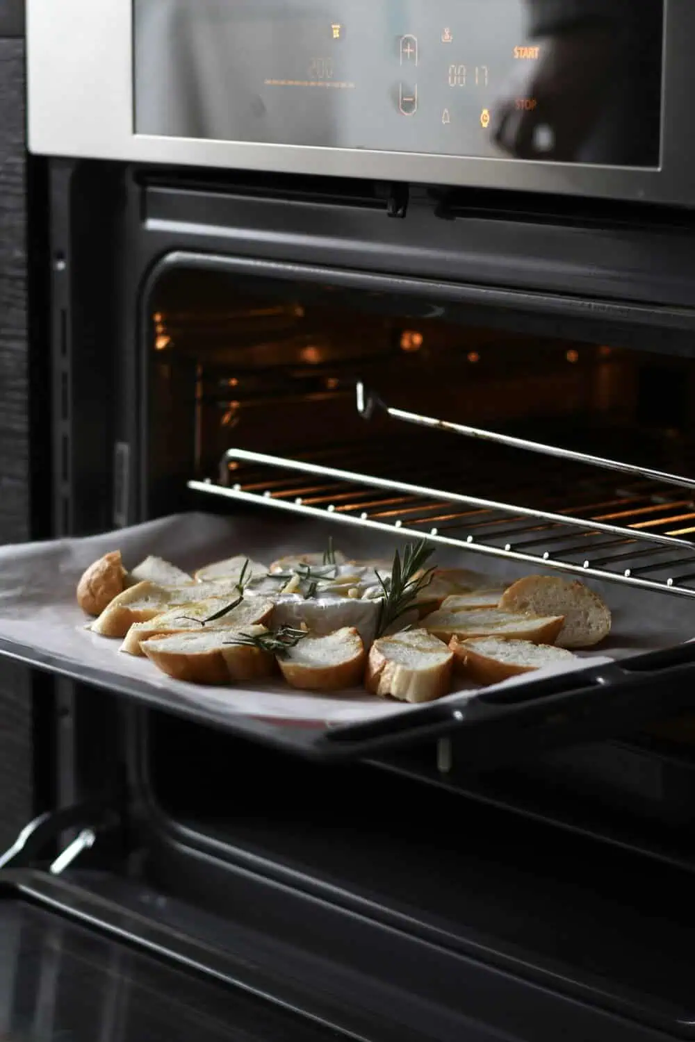 An oven rack is pulled fully out holding a tray of toasted baguette slices with melted cheese and fresh rosemary, with the oven's digital display visible above.