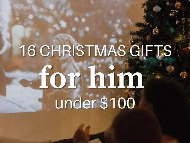 A close-up crop of a movie projected onto a wall beside a decorated Christmas tree, with two people watching from a couch, overlaid with text reading "16 Christmas gifts for him, under $100."