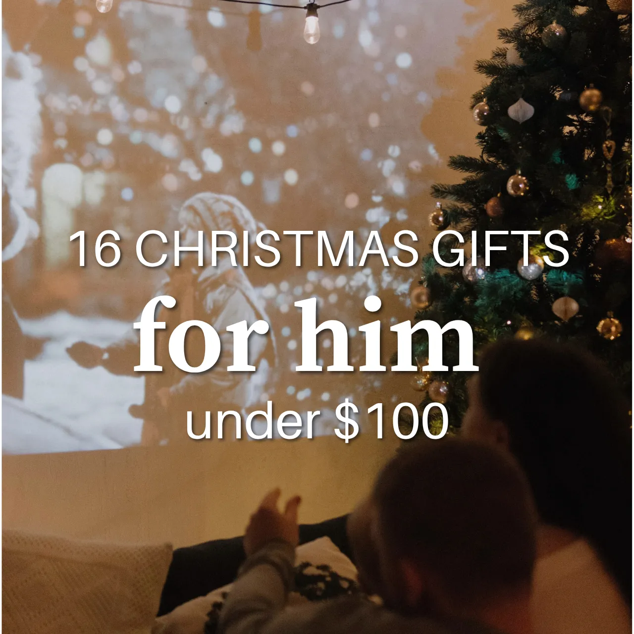 A movie projects onto a wall beside a decorated Christmas tree strung with string lights, with two people lying on couch cushions watching, overlaid with text reading "16 Christmas gifts for him, under $100."