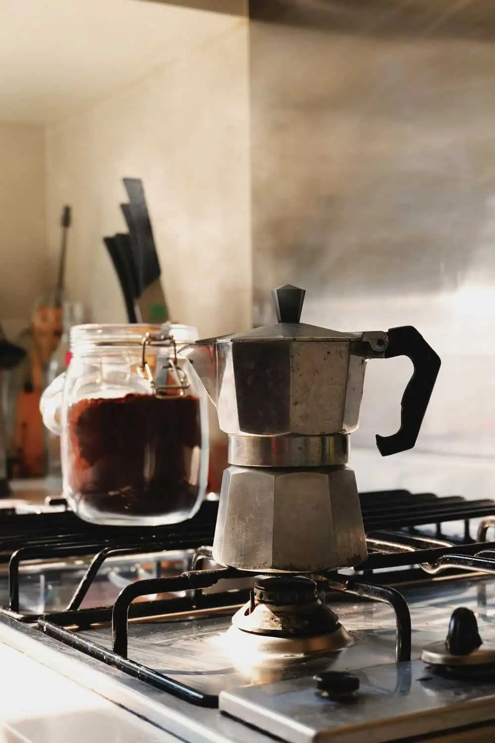 A stovetop moka pot with a black handle sits on a gas burner next to a glass jar of dark roast coffee, backlit by warm morning light.