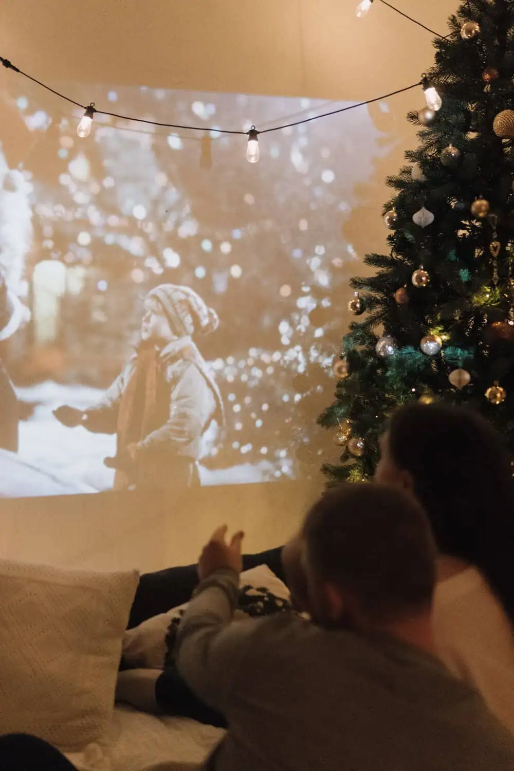 A movie projects onto a wall beside a decorated Christmas tree strung with string lights, with two people lying on couch cushions watching.