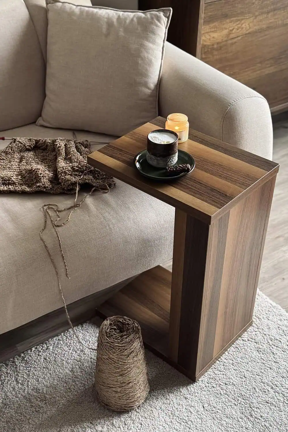 A wood C-shaped tray table beside an armchair holds a lit candle and a mug on a saucer, with a chunky knit blanket and a ball of twine nearby.