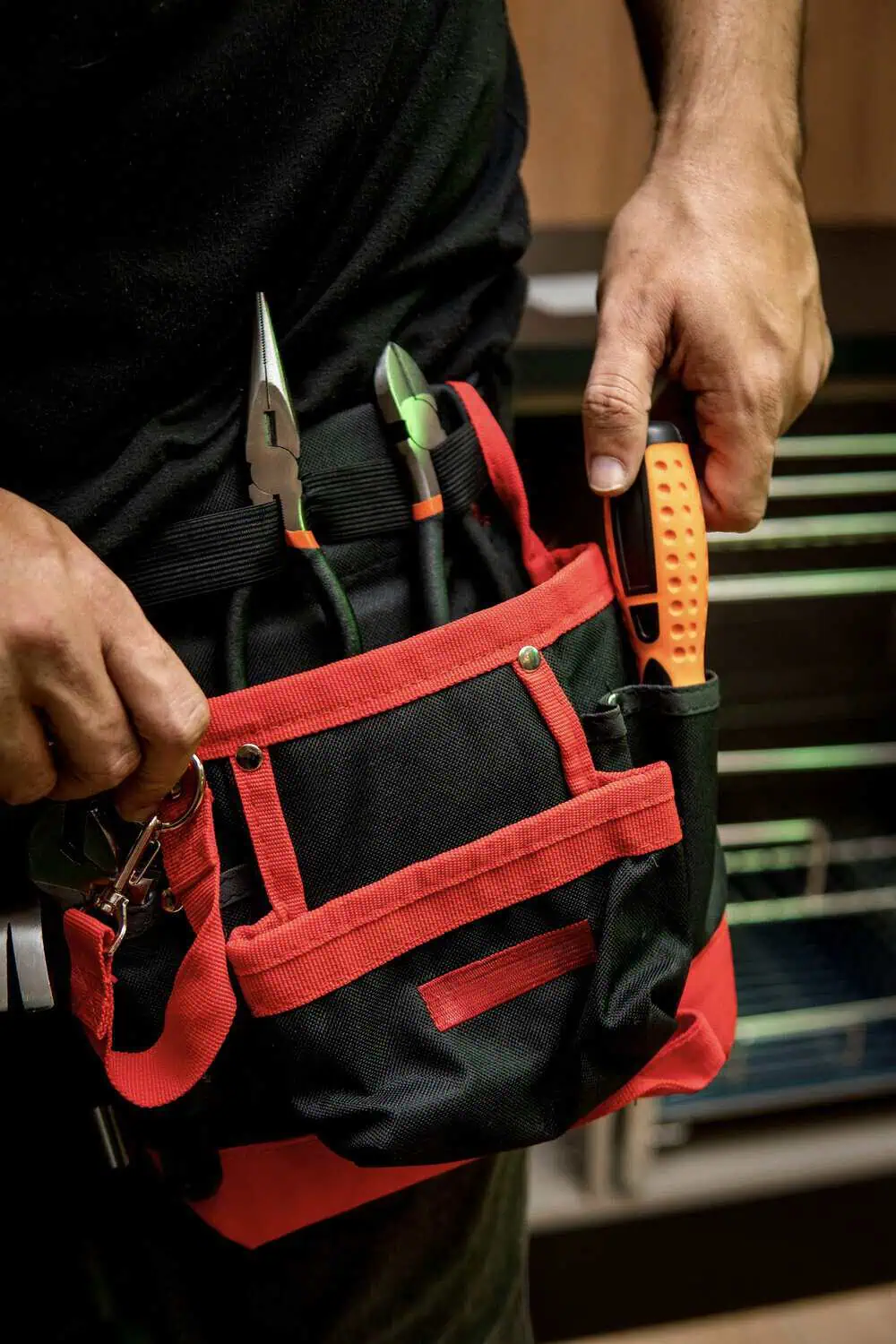 A man holds a red and black zippered tool bag loaded with pliers and other hand tools, standing in front of a workbench.