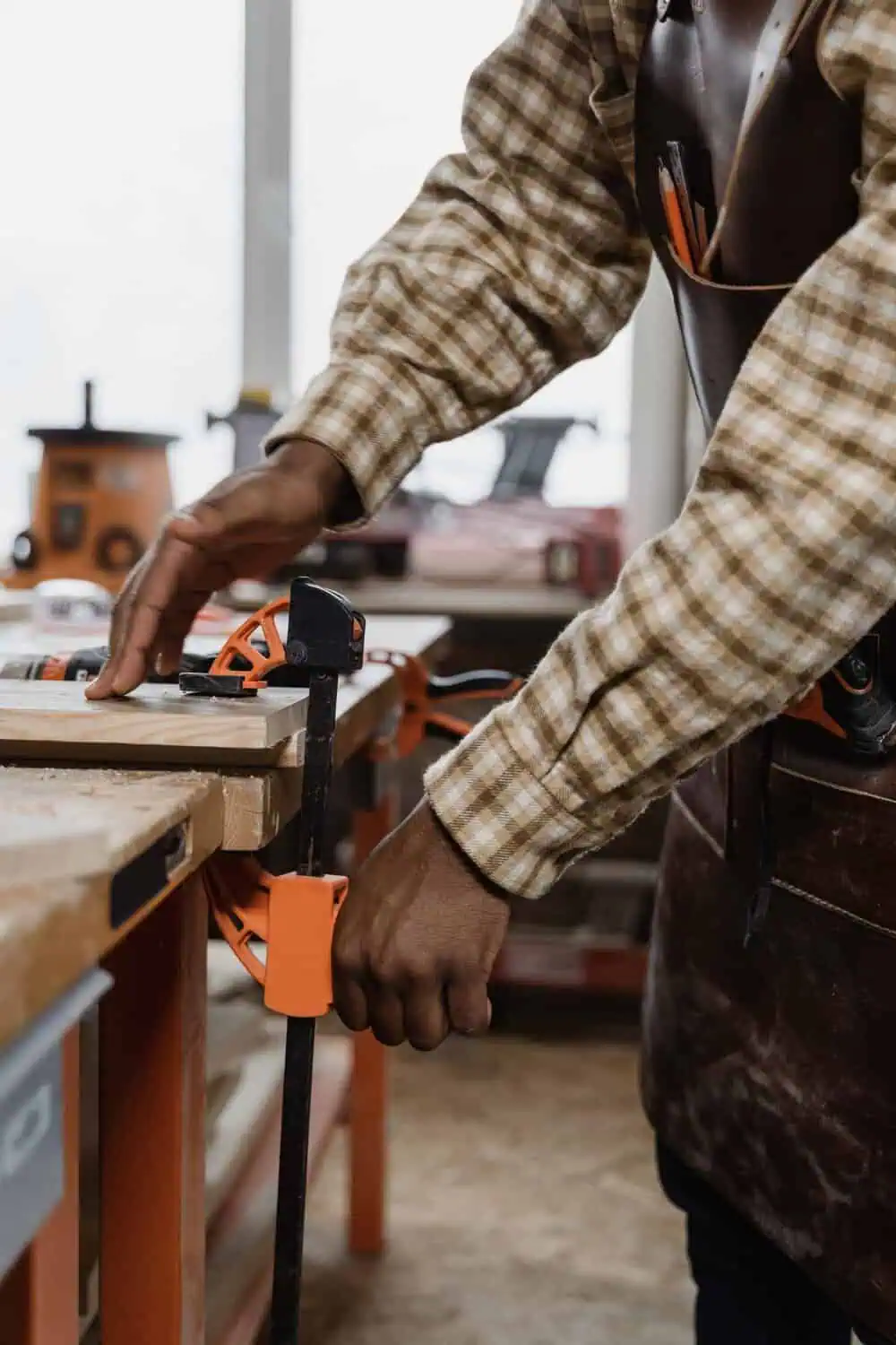A man in a leather apron tightens an orange bar clamp holding a piece of wood on a workbench, with a pegboard of tools in the background.
