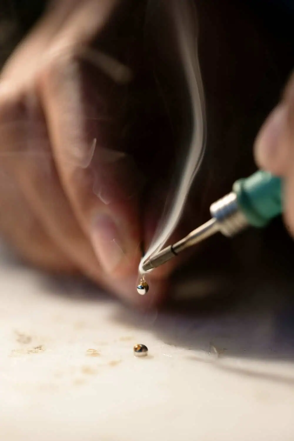 A soldering iron tip melts a bead of solder with a thin curl of smoke rising above a workbench surface.