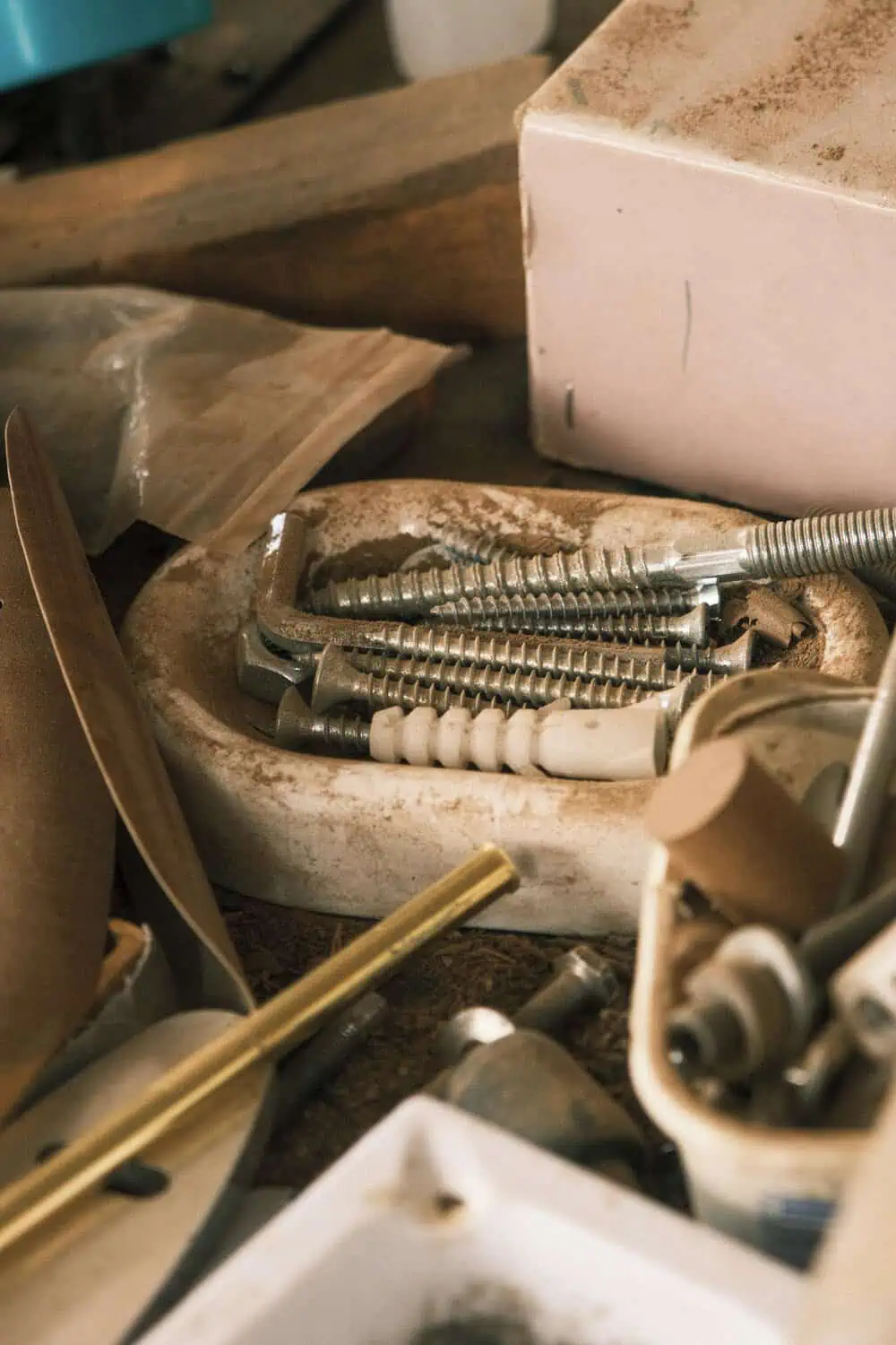 A ceramic bowl holds an assortment of loose screws and hardware pieces, surrounded by workshop clutter including a folded paper sleeve and a small cardboard box.