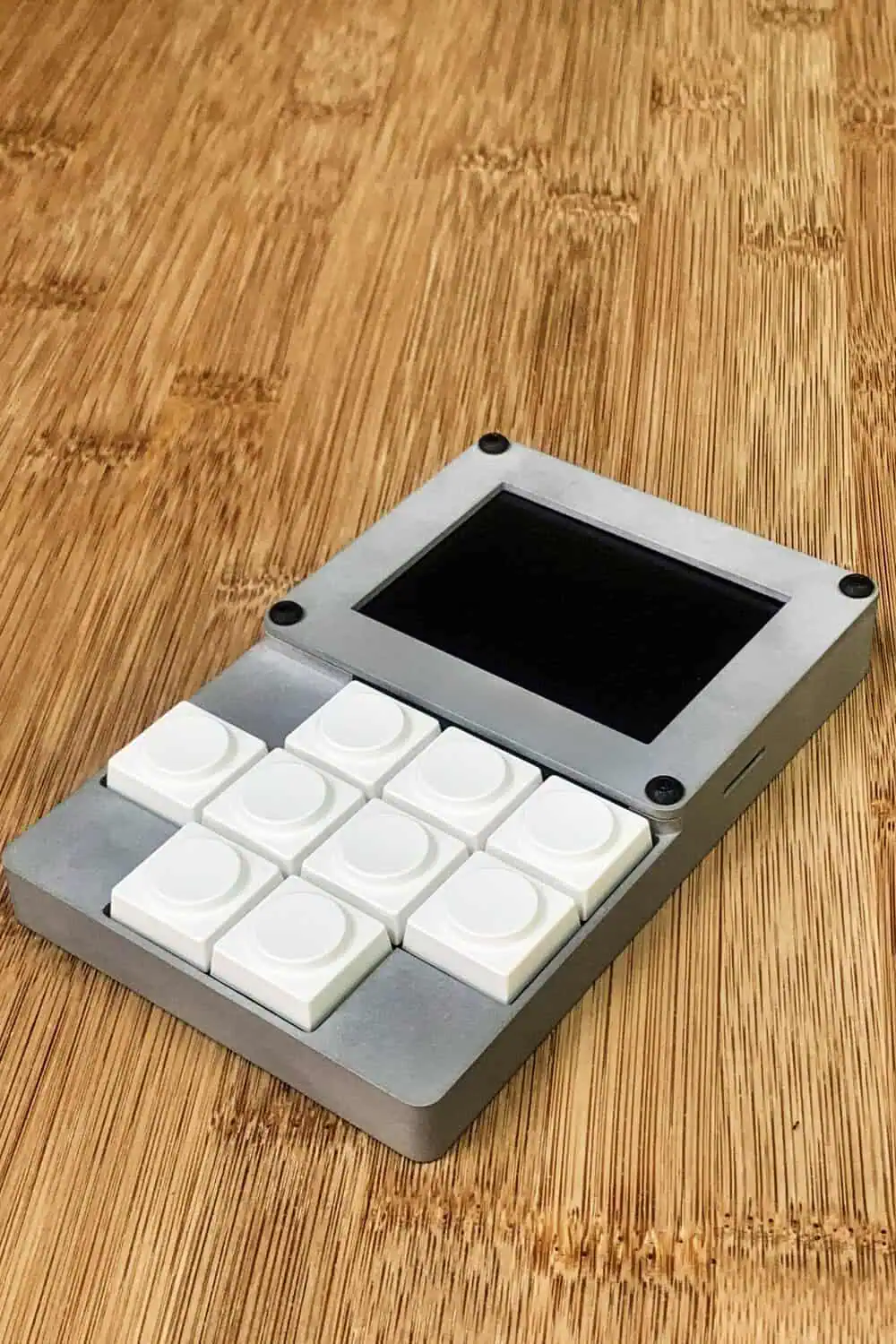 A gray macro keypad with eight white buttons and a small screen sits at an angle on a wood desk.