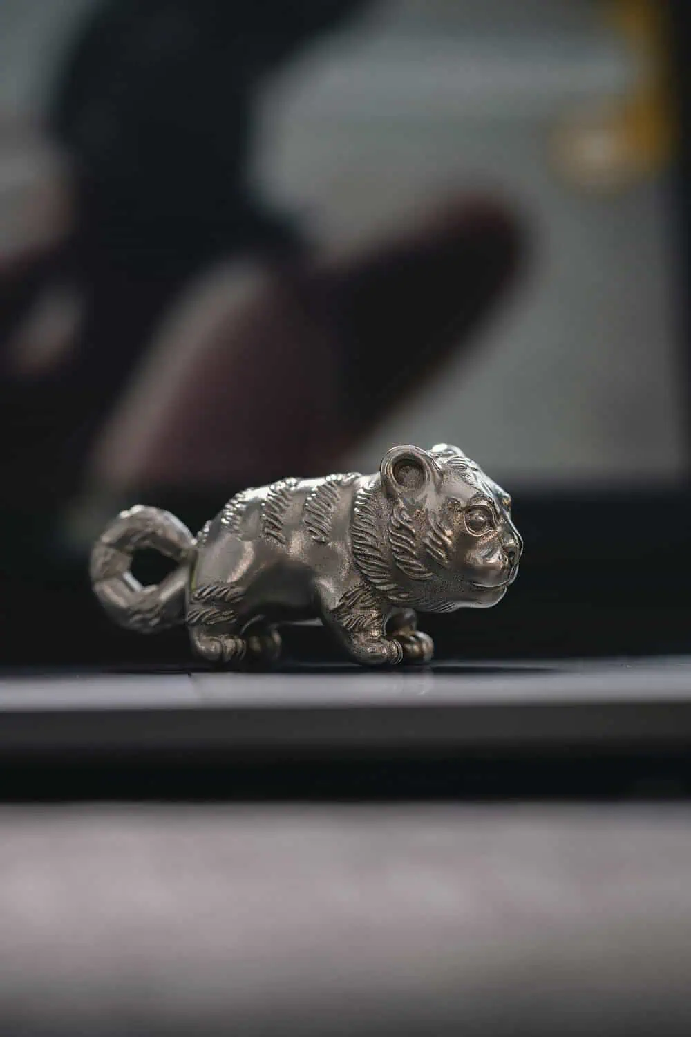A small silver metal figurine of a crouching tiger cub sits on a dark reflective surface.