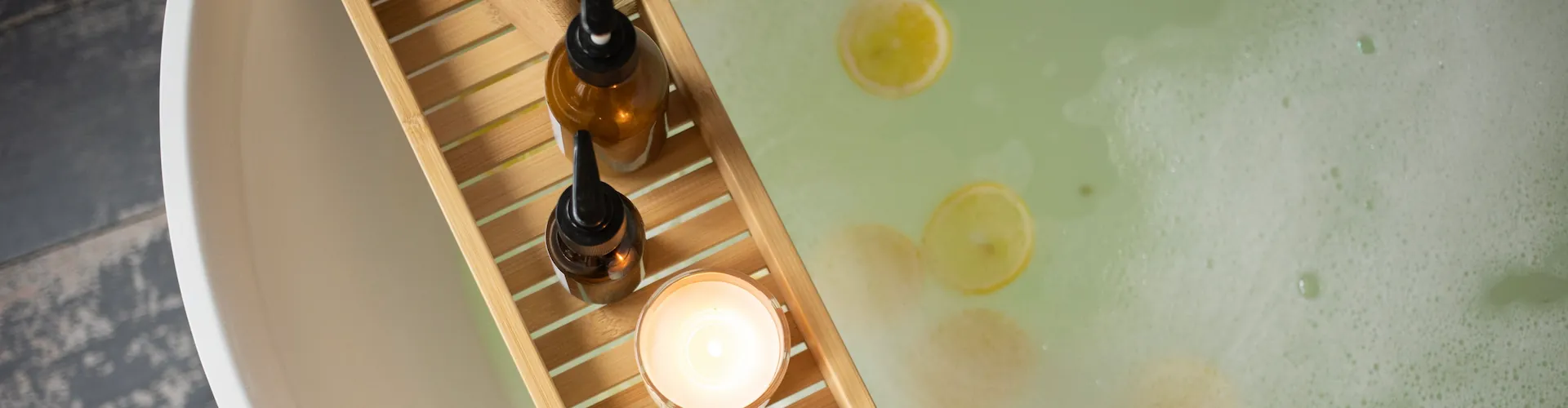 A bamboo bath tray with oil bottles and a lit candle over a lemon-scented bath.