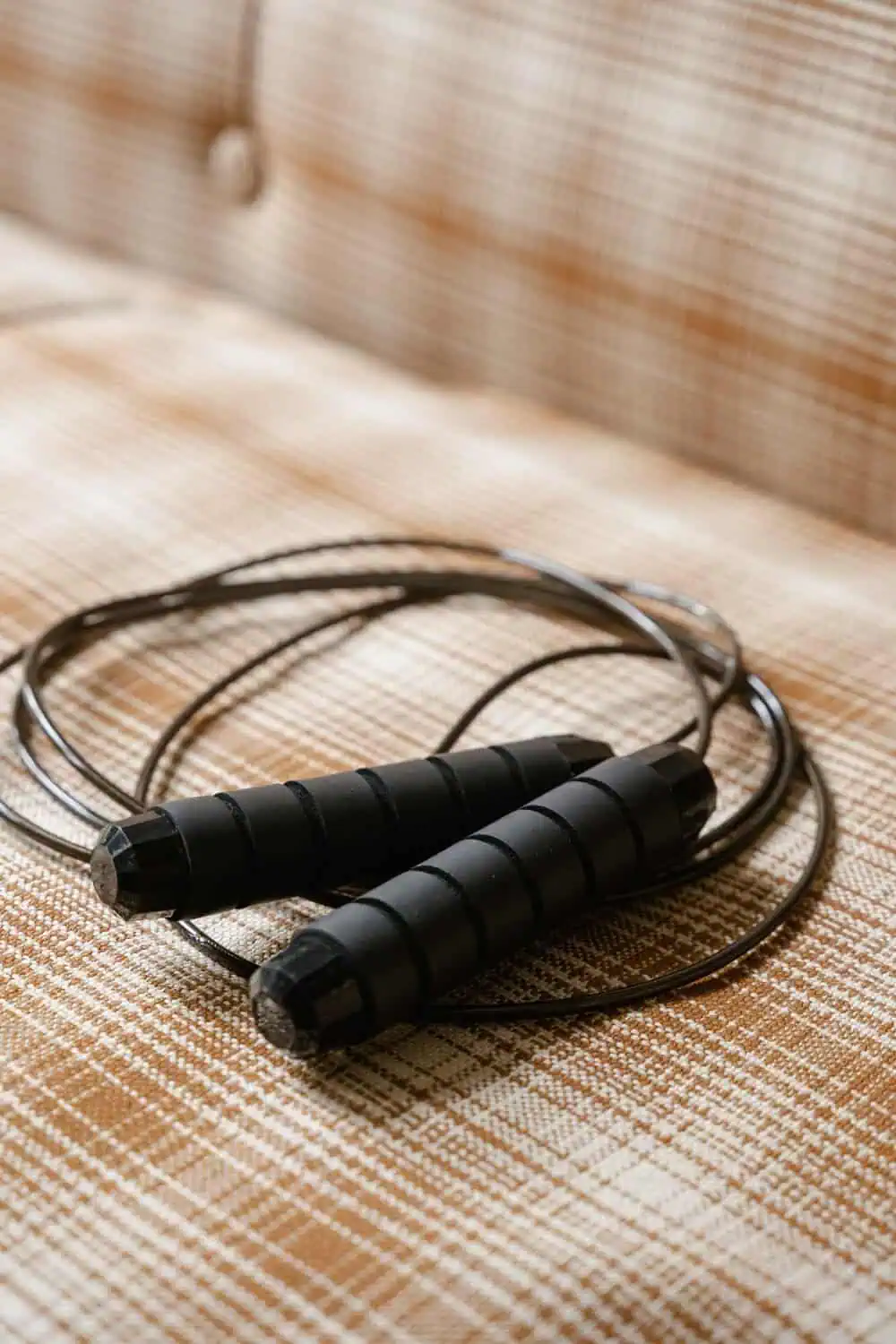 A coiled jump rope with black weighted handles resting on a woven cushion.