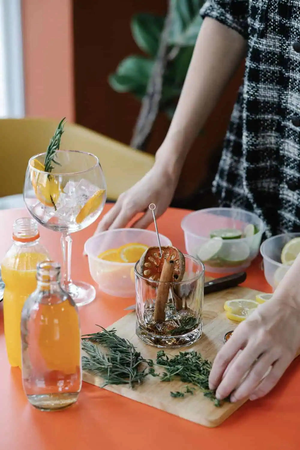 Hands preparing a garnished cocktail with citrus and herbs on a wooden board.