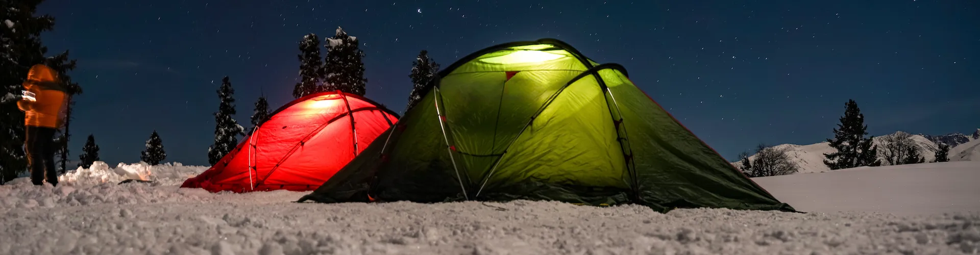 Two illuminated tents, one red and one green, glow at night in a snowy campsite under a starry sky.