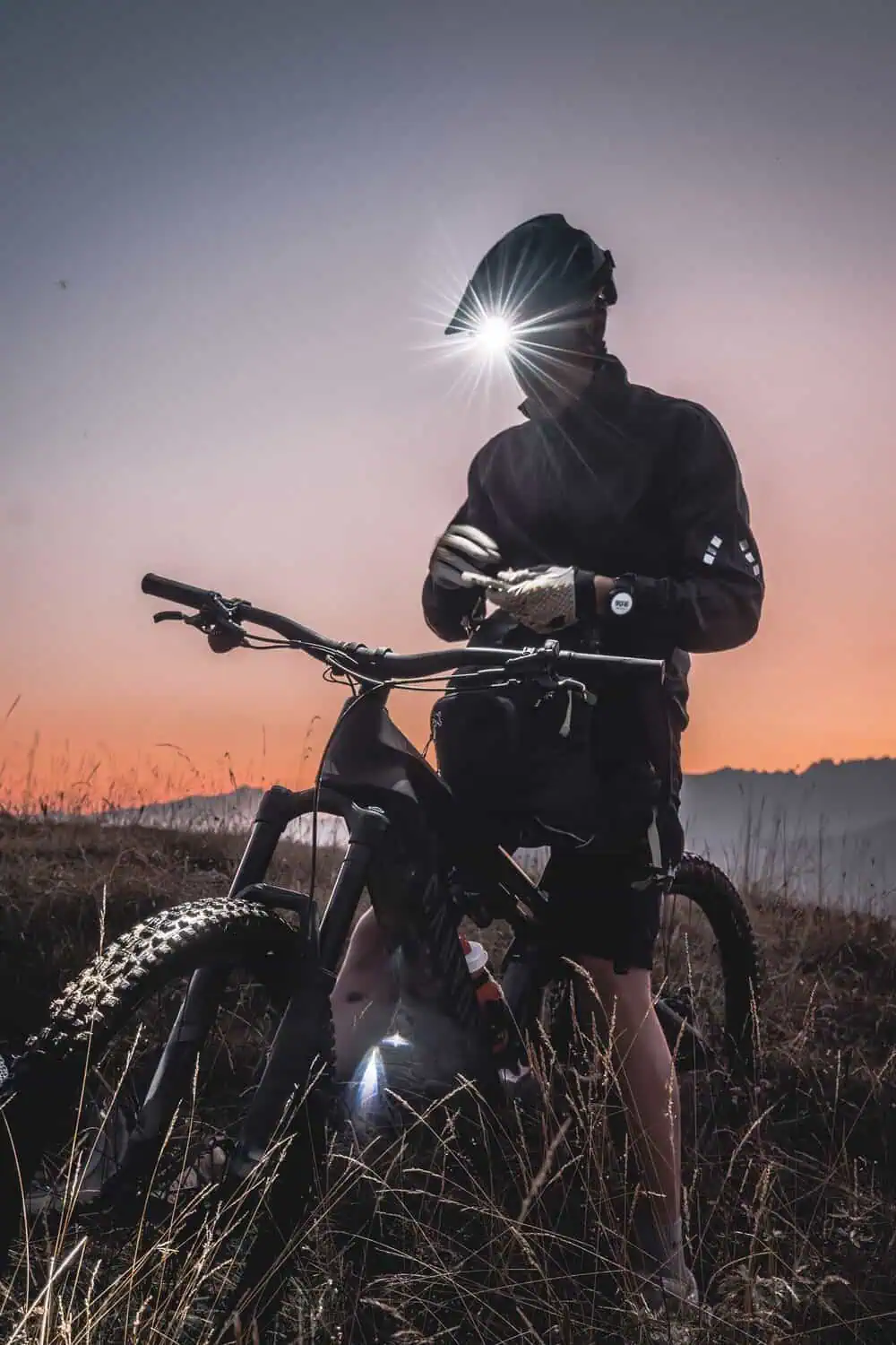 A cyclist stands with a mountain bike at dusk, a bright headlamp beam glowing against the darkening sky.