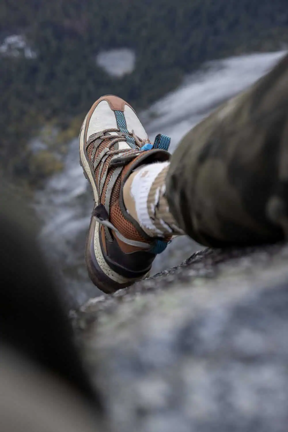 A close-up of a hiking boot and thick wool sock resting on a rock ledge with a forest below.