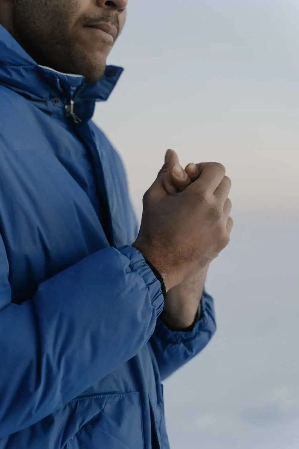 A person in a blue puffer jacket clasps their hands together outdoors against a hazy sky.