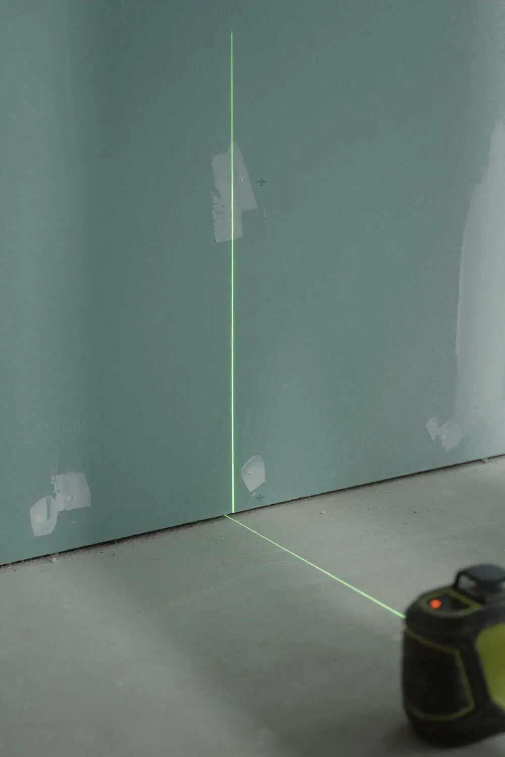 A laser level projects a bright green line up a wall corner from a small device on the floor.