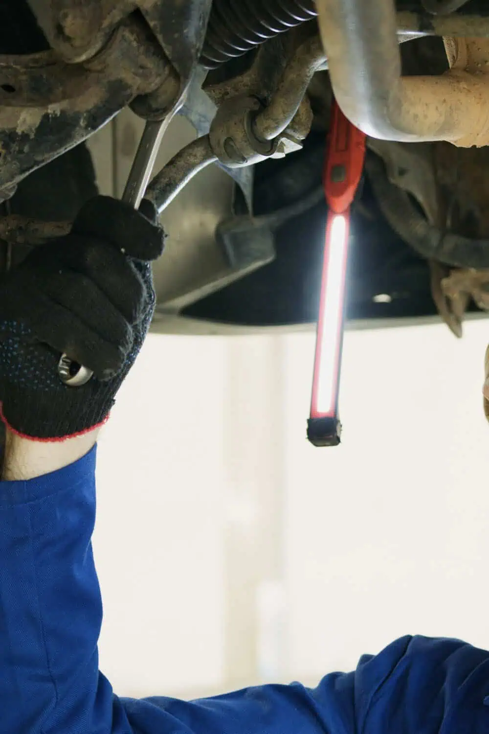 A mechanic's gloved hand works a wrench under a car, lit by a red cordless work light clipped nearby.