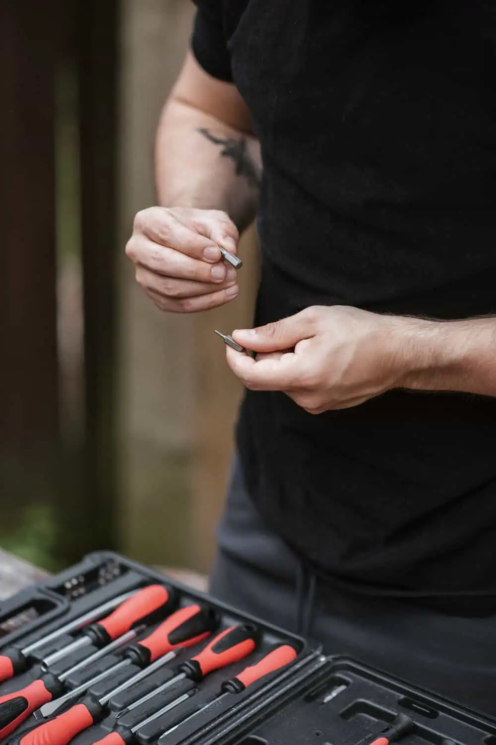 A pair of hands holds two interchangeable screwdriver bits above an open case of red-handled screwdrivers.