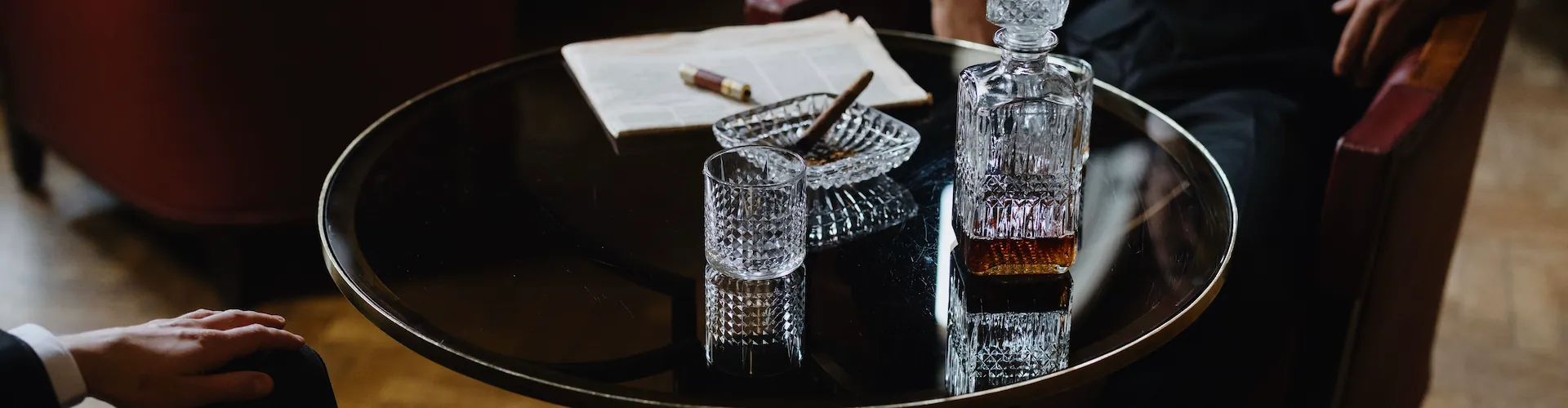 A round table holds a whiskey decanter, a glass, a lit cigar resting in an ashtray, and a folded newspaper.
