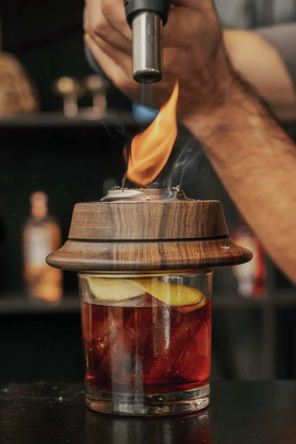 A torch flame rises above a wooden smoking dome covering a cocktail glass with citrus and ice.