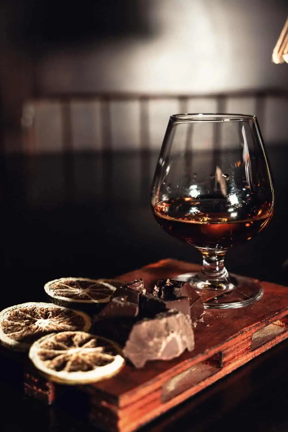 A snifter of brandy sits next to chunks of dark chocolate and dried citrus slices on a wood board.
