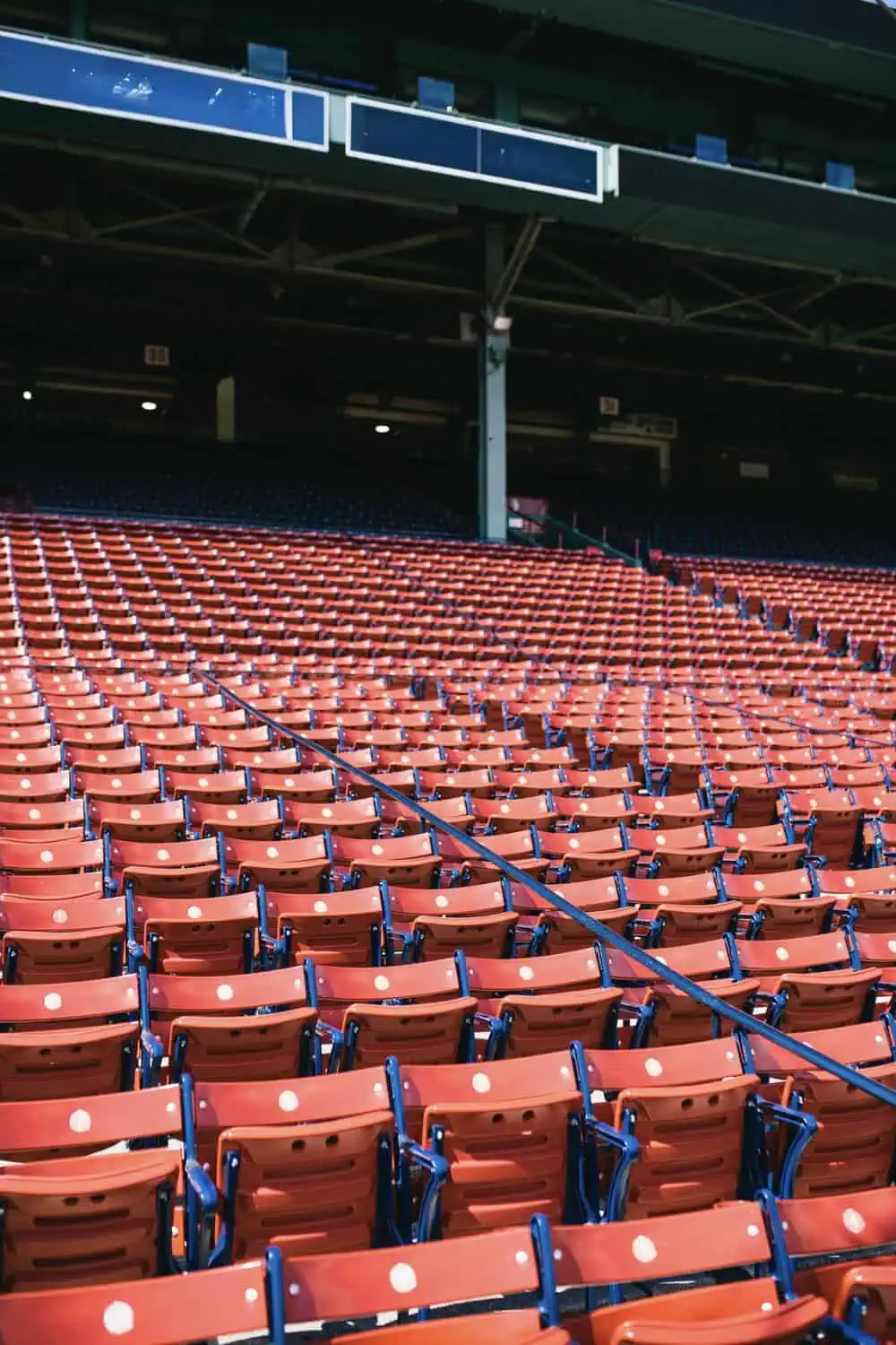 Rows of empty orange stadium seats rise toward a covered upper deck under stadium lighting.